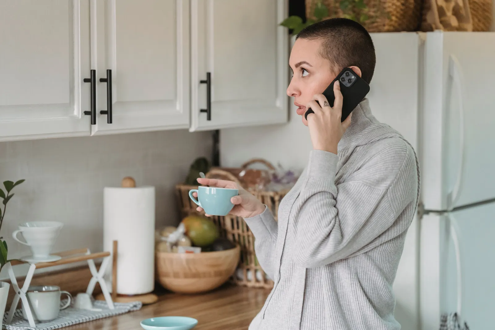 A woman standing in her kitchen holding a mug while talking on her phone