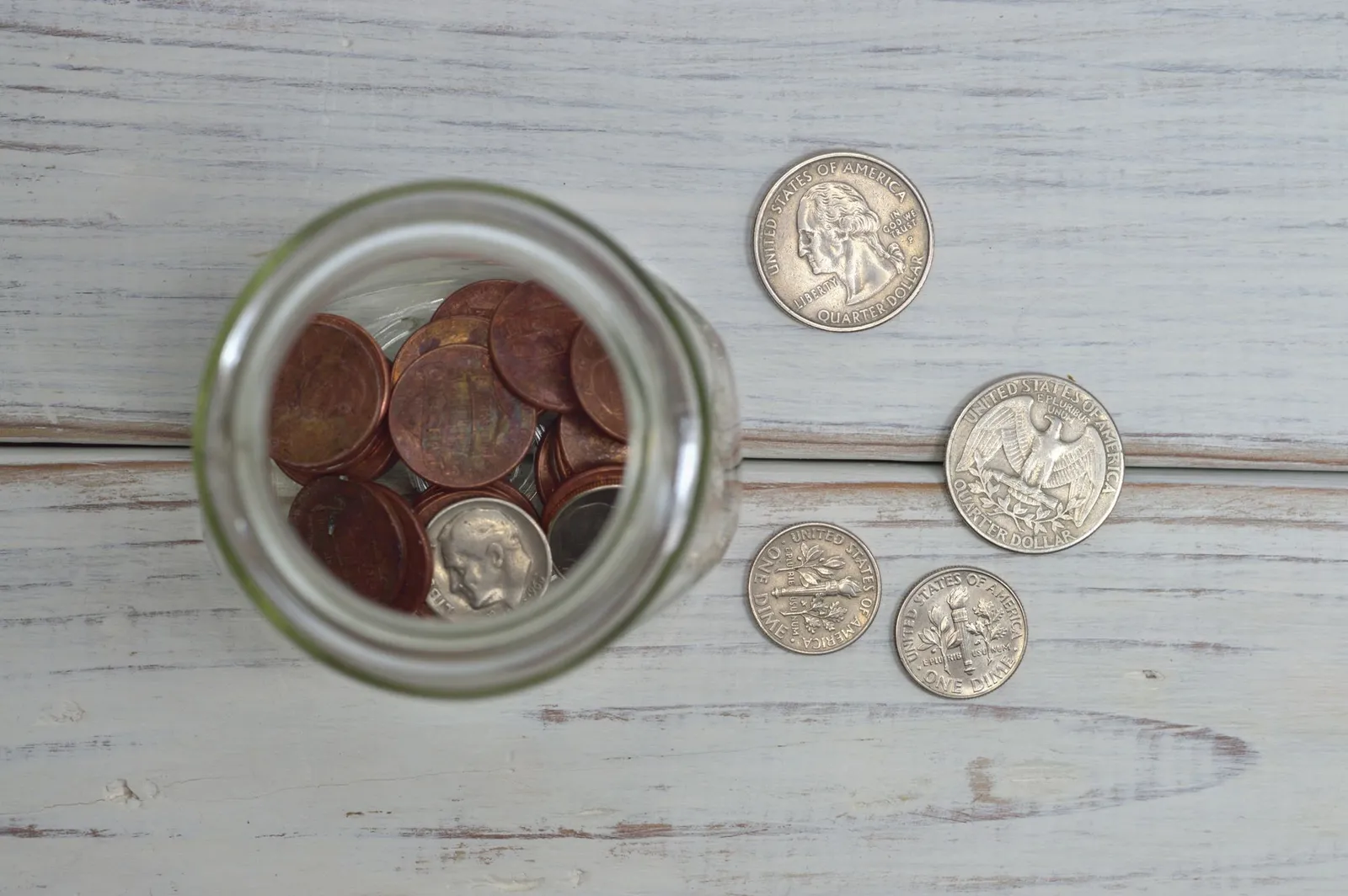 Top-down view of a glass jar of pennies next to two quarters and two dimes on a whitewashed wooden table