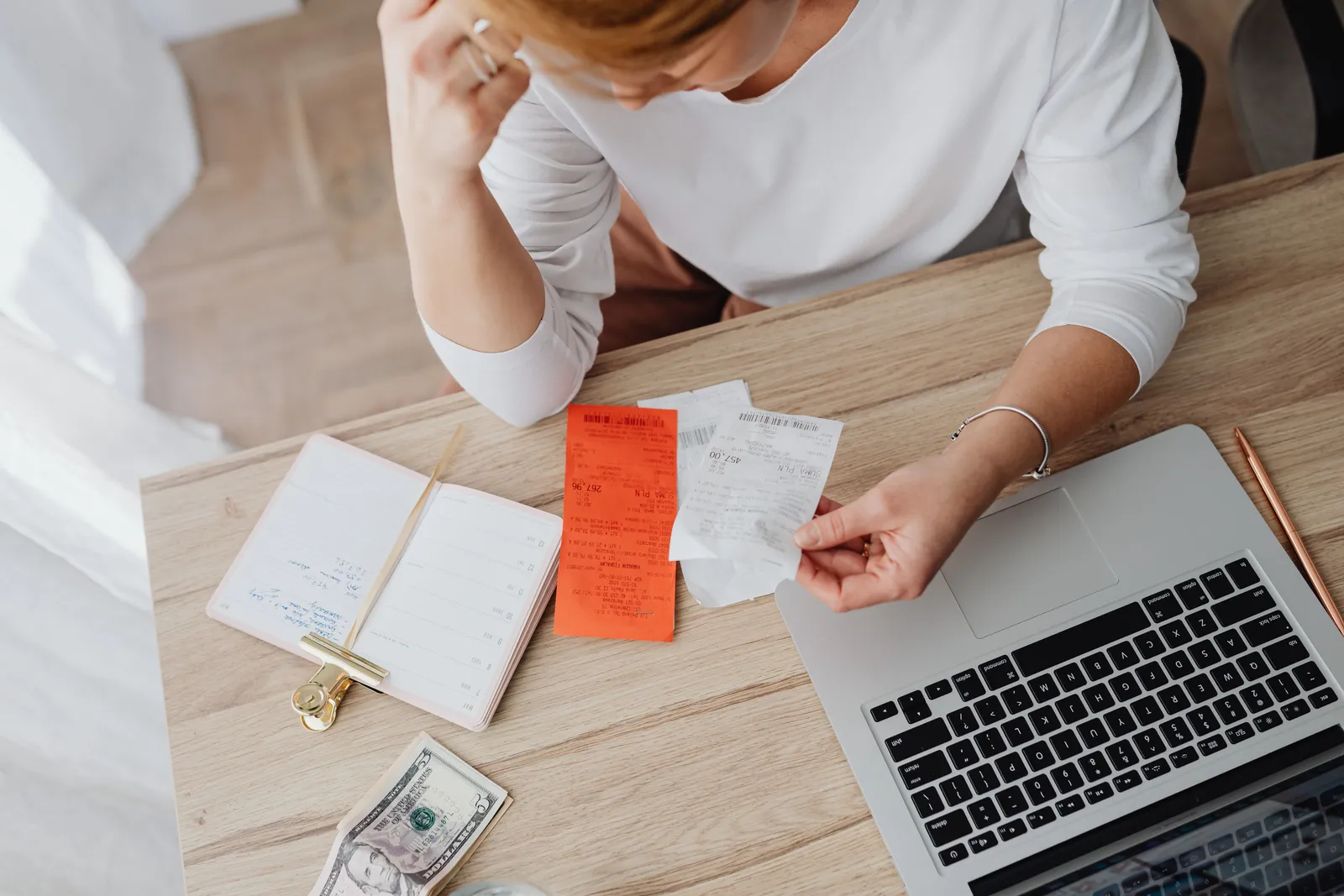 A woman at a desk sorting paper receipts beside a laptop, an open notebook, and cash