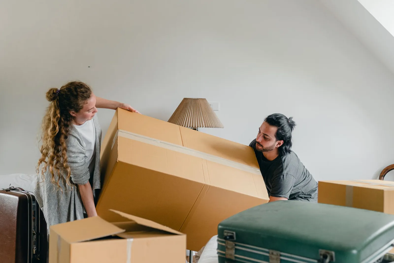 A young couple lifts a large moving box in a bright attic room surrounded by suitcases
