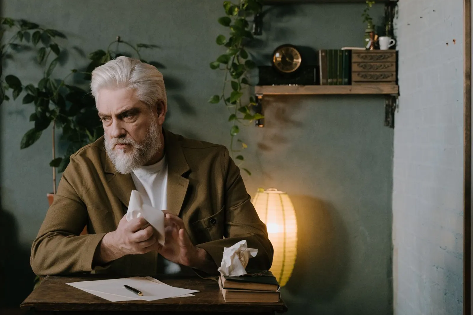 A gray-haired man at a wooden desk crumpling a sheet of paper, blank pages and a pen in front of him