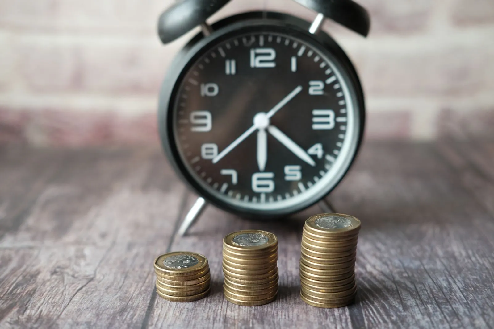 Black alarm clock behind three short stacks of coins on a wooden table