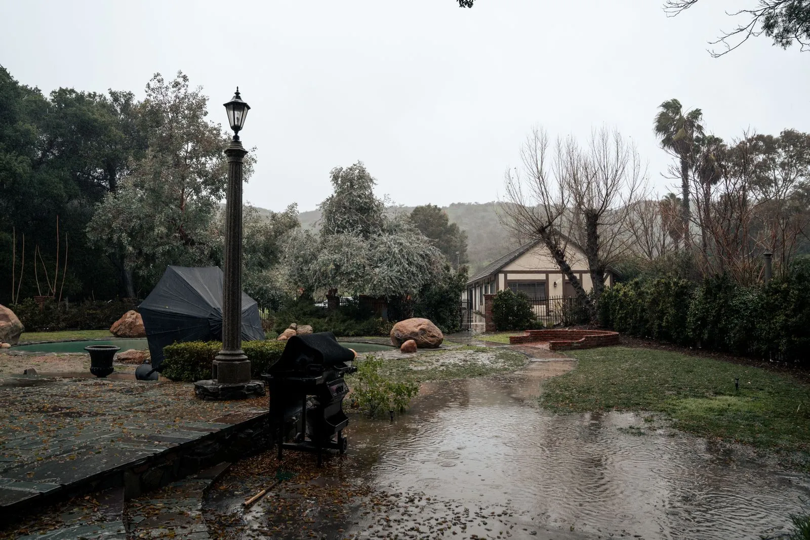 Rainwater pooling across a backyard lawn and patio at a hillside home with palm trees during a storm
