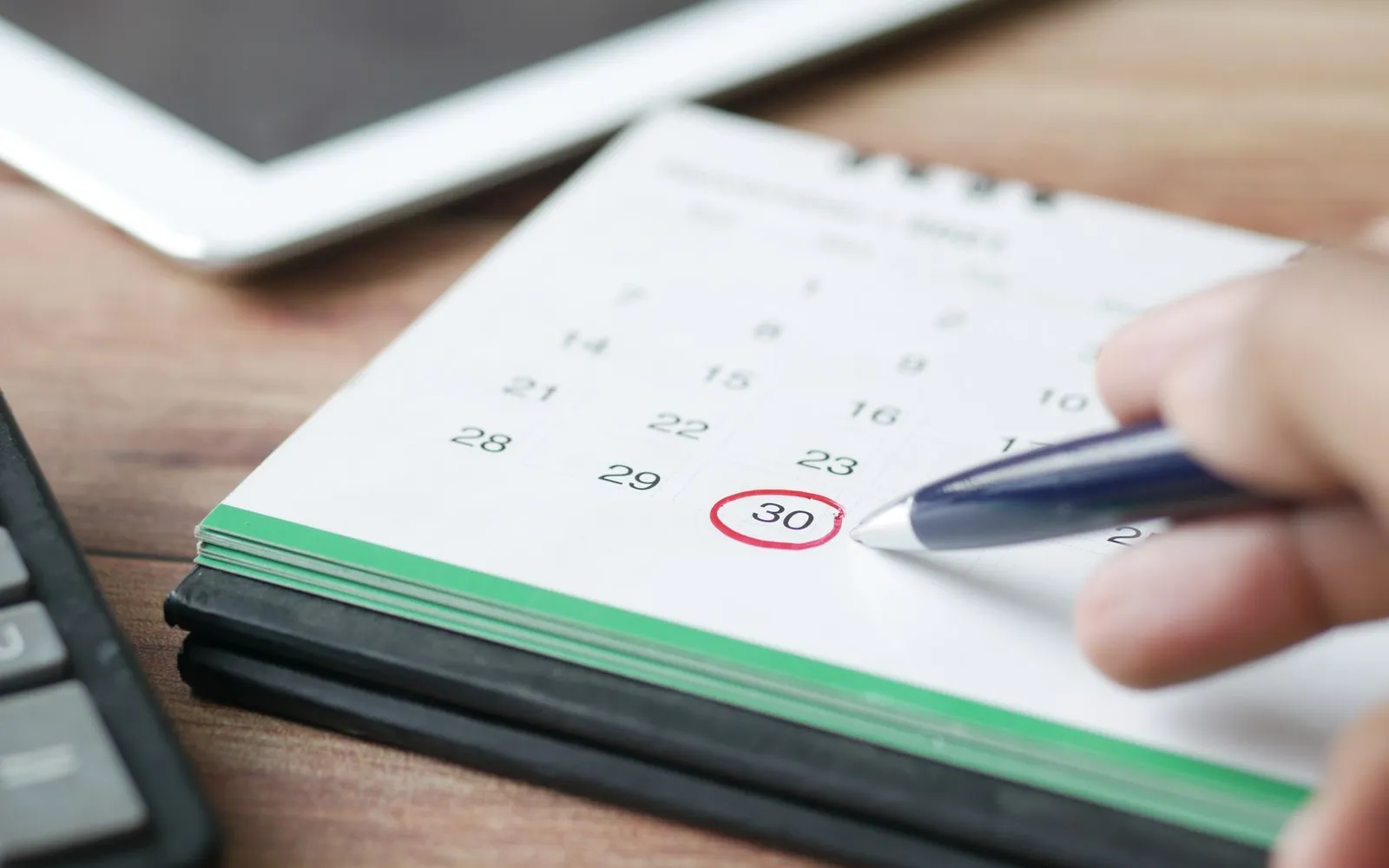 A hand marking a date on a paper wall calendar with a pen