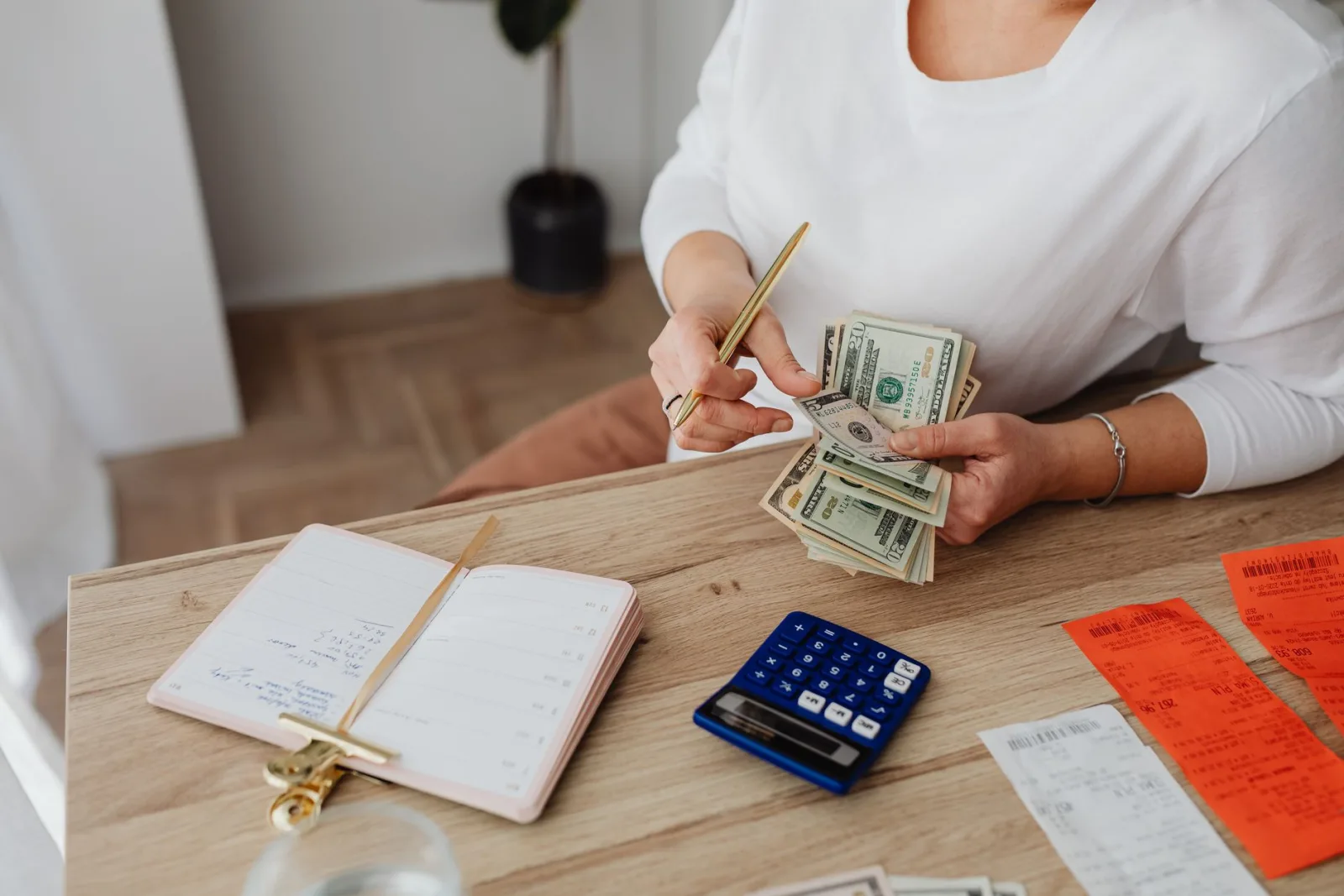A woman at a wooden desk counting cash beside a calculator, a notebook and a stack of receipts