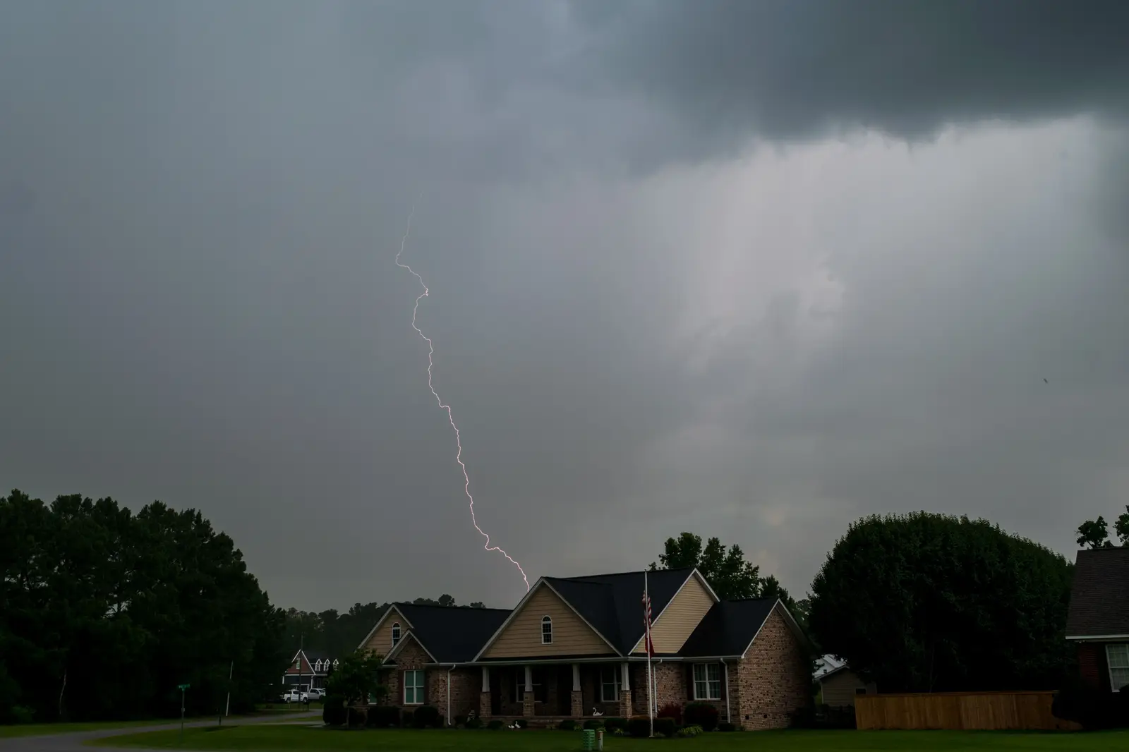 Lightning over a brick house with a flagpole under dark storm clouds