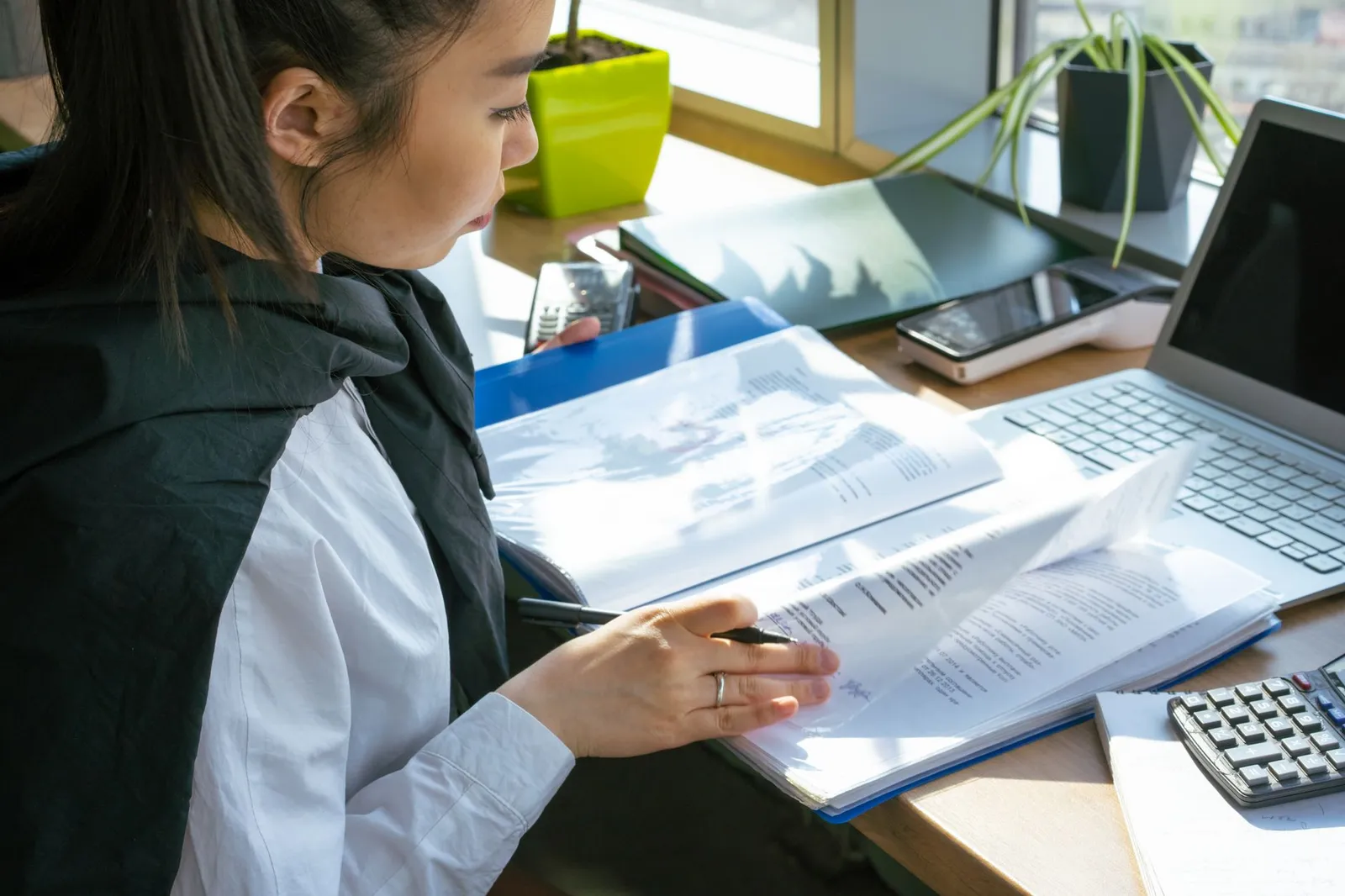 A young woman with a pen going through a folder of documents beside a laptop and a calculator