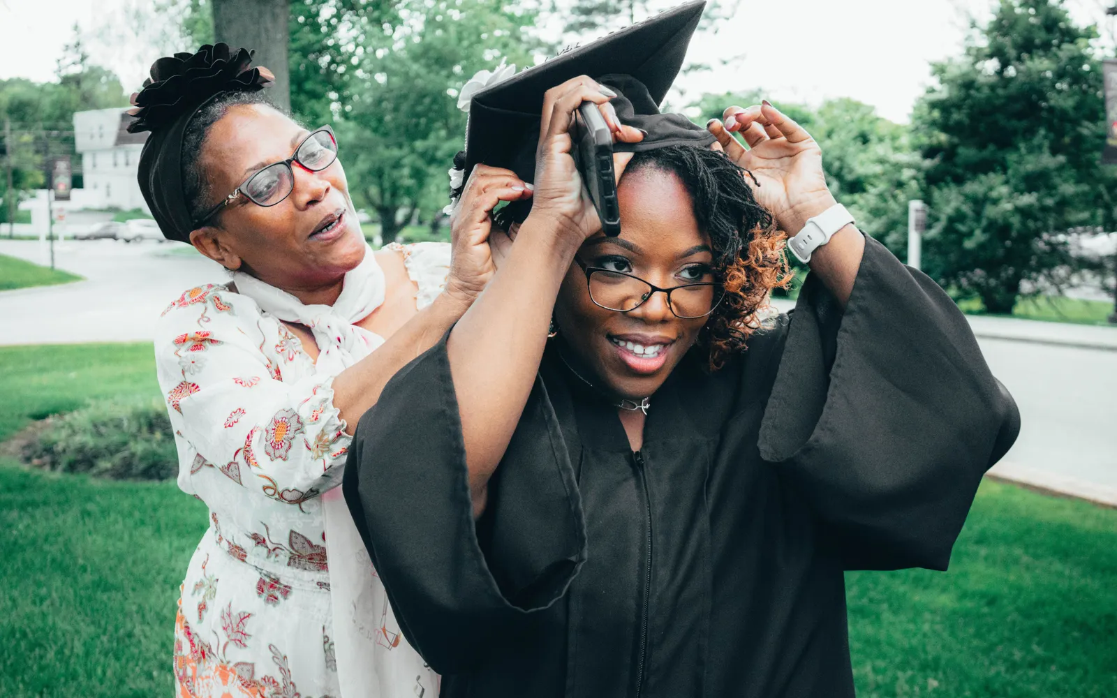 A mother adjusts her daughter's graduation cap outdoors on a green campus lawn