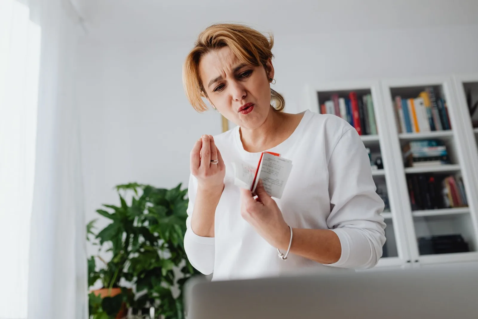 A woman frowns at a folded paper receipt while standing at a desk in a bright room