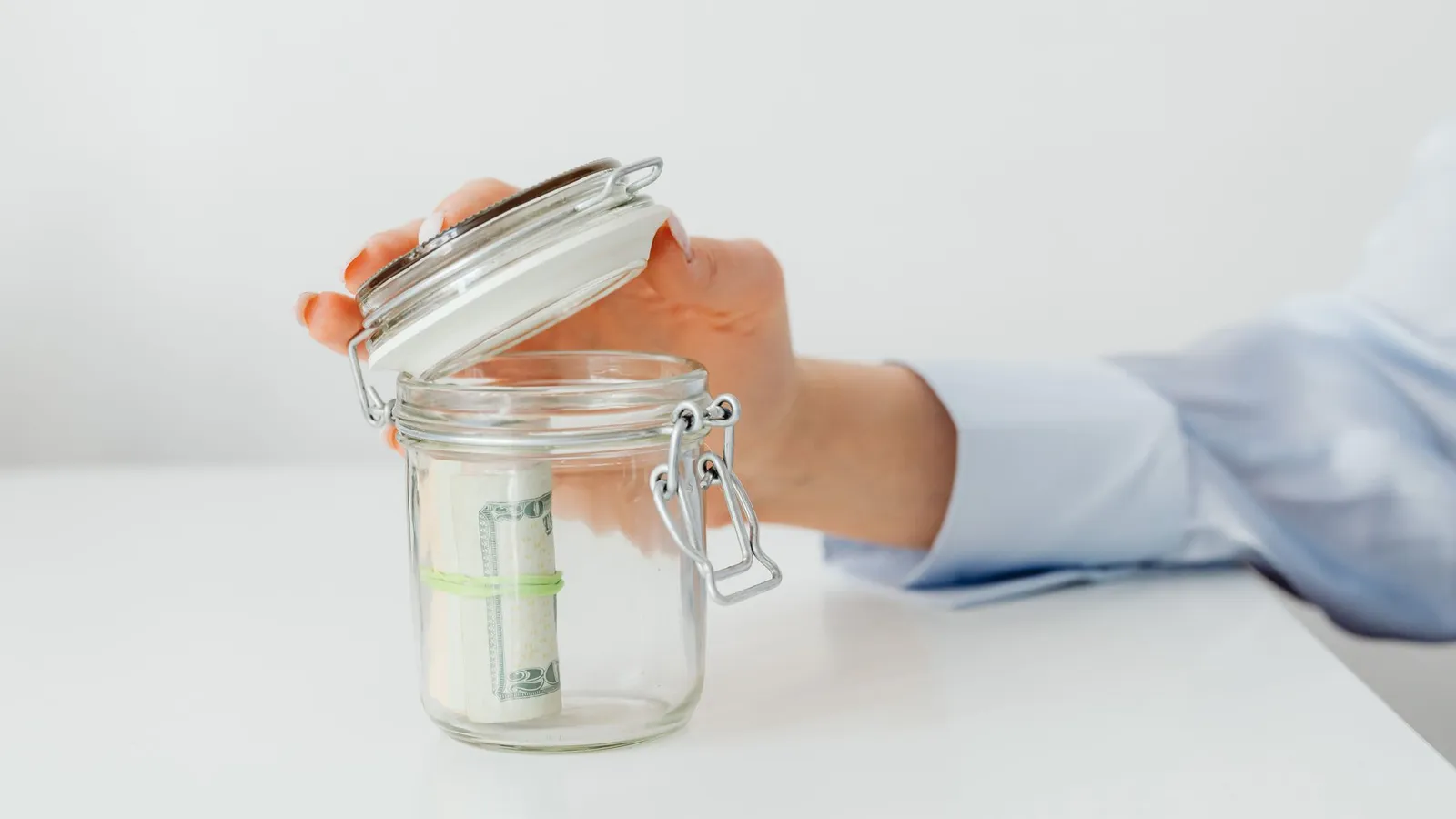 A hand lifting the lid of a glass jar holding a rolled stack of dollar bills on a white table