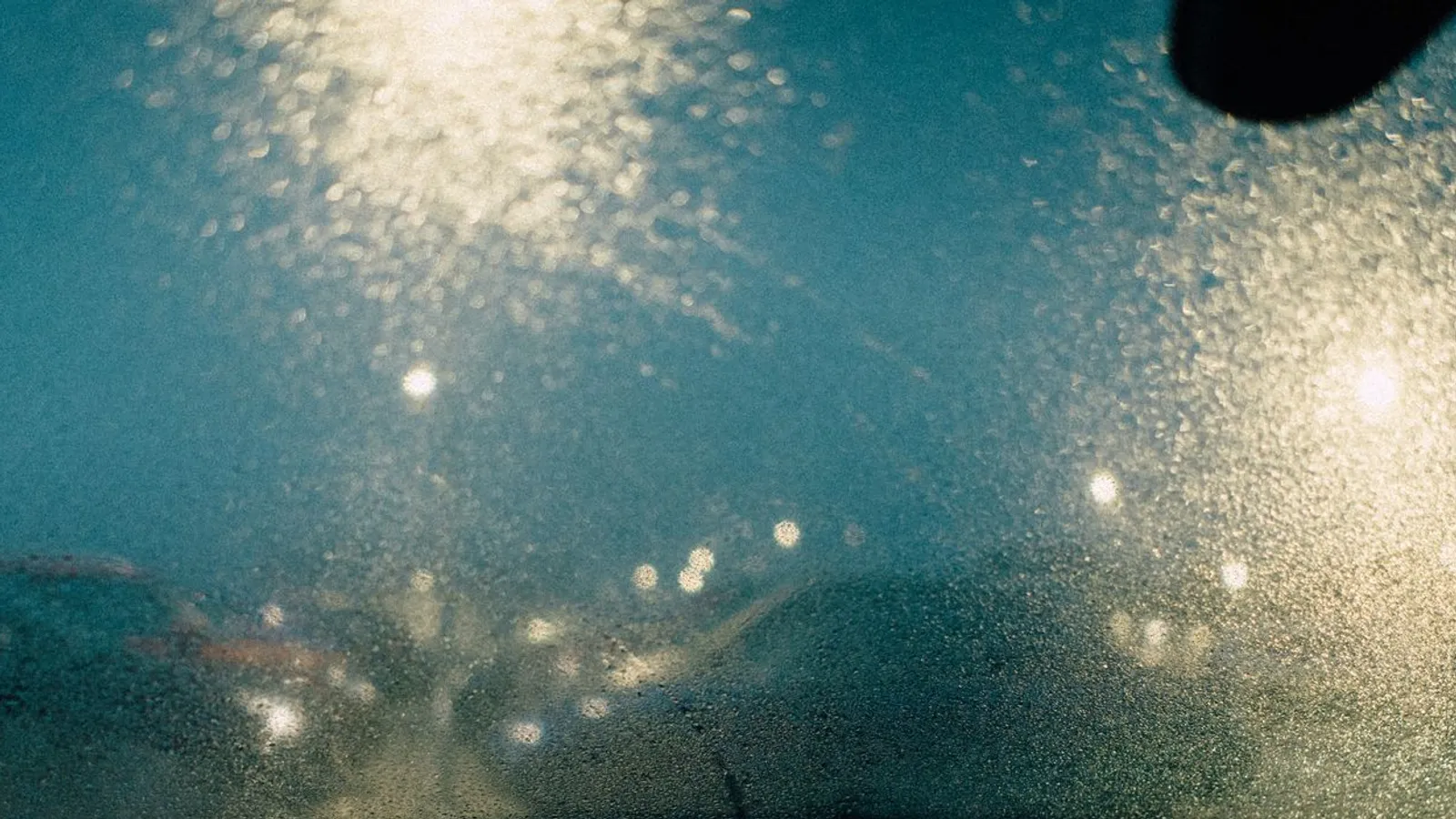 Rain-covered windshield at dusk with blurred headlights and streetlights beyond the glass