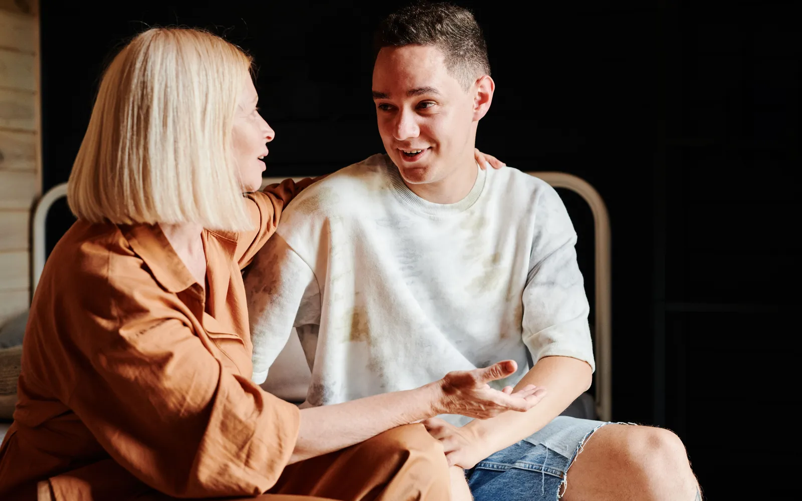 A mother talks with her college-age son as they sit side by side on a bed