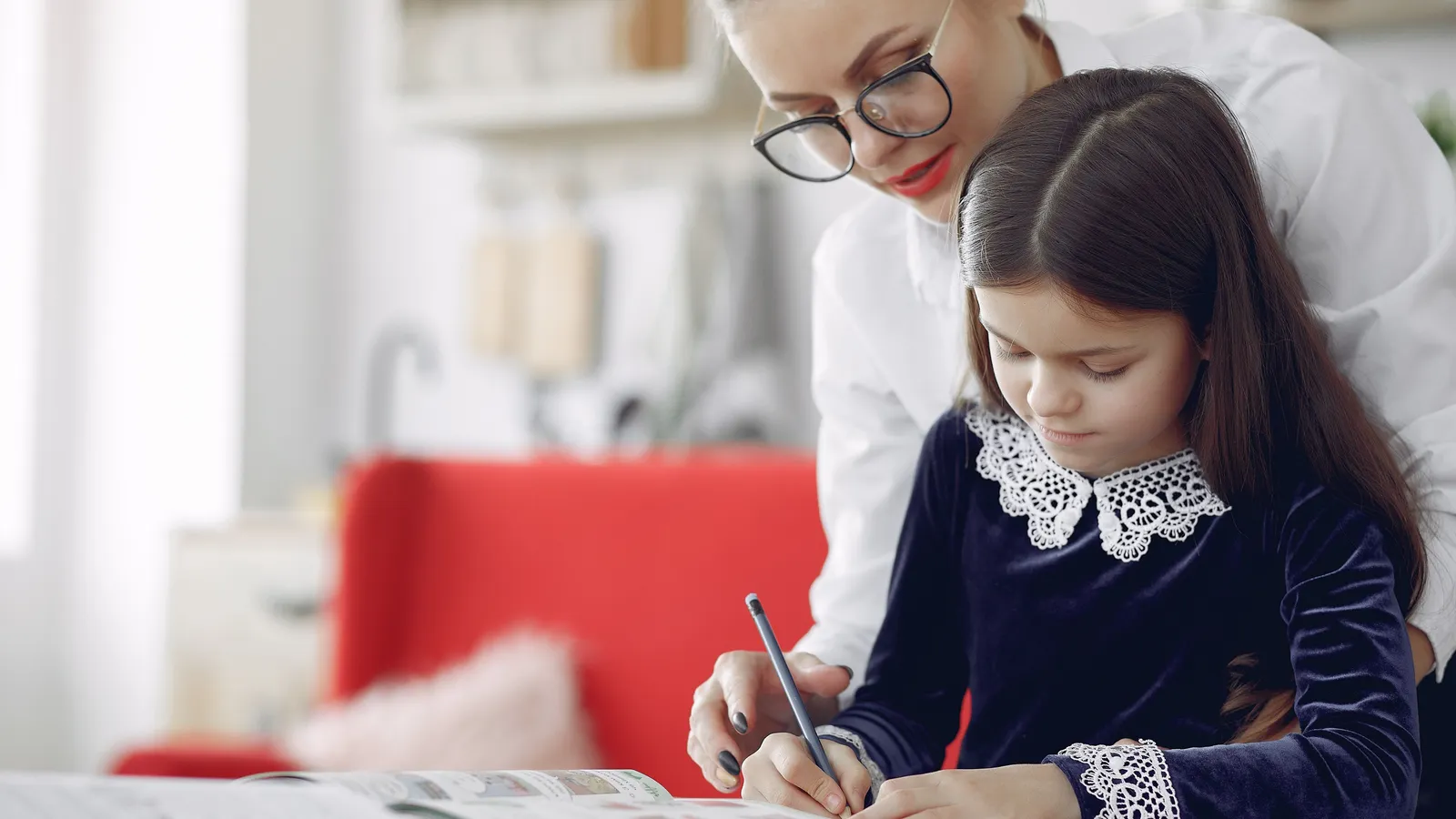 A tutor in glasses leaning over a girl as she writes in a workbook at a table