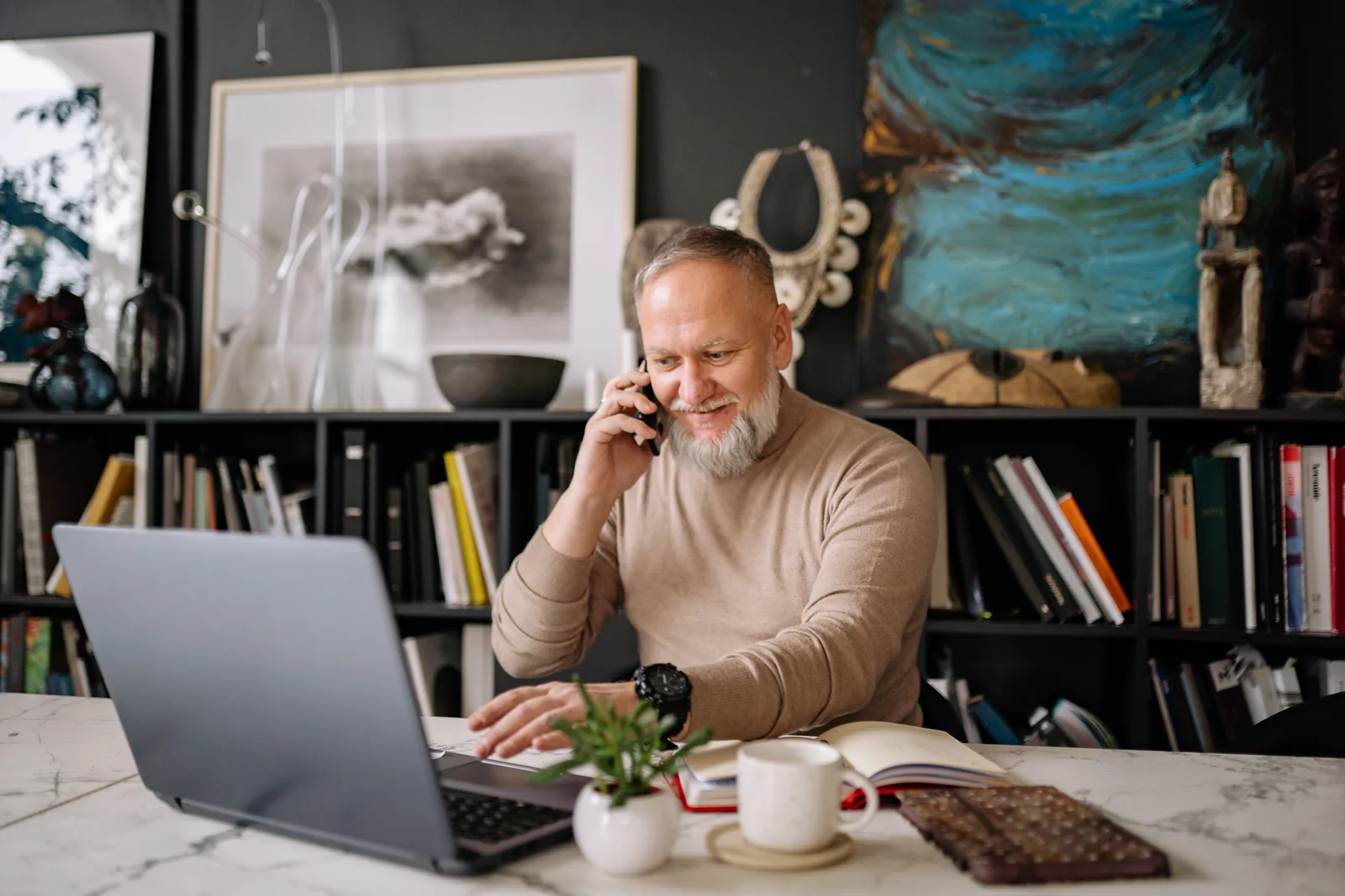 An older man working on a laptop at home while taking a phone call