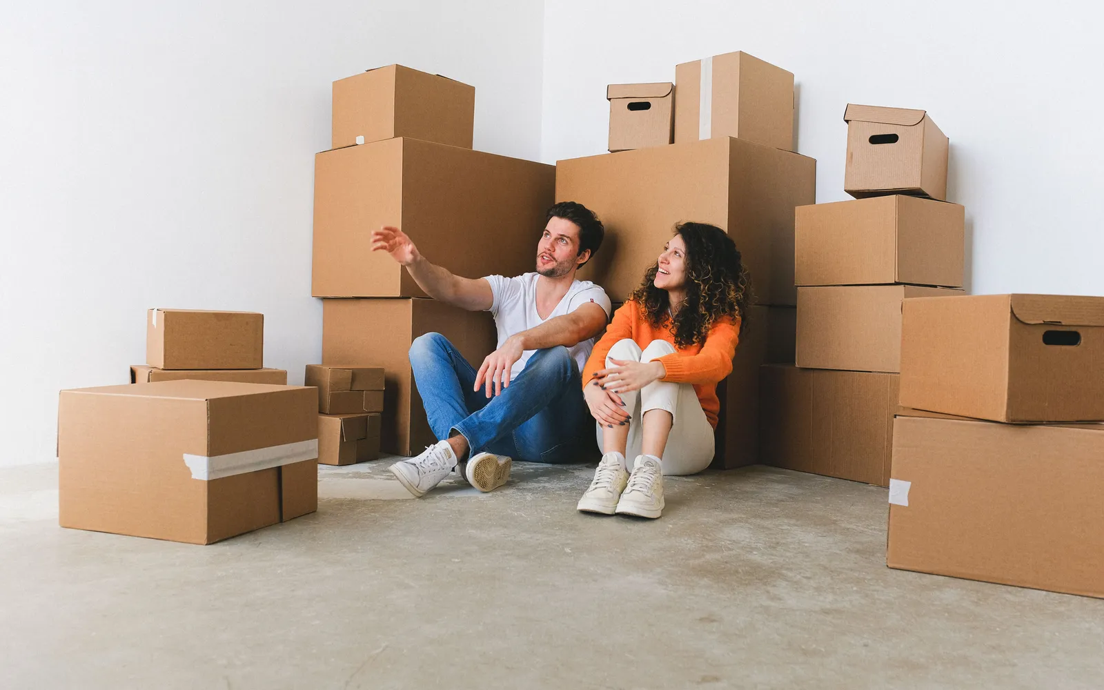 A young couple sitting on the floor among stacked moving boxes in an empty white room