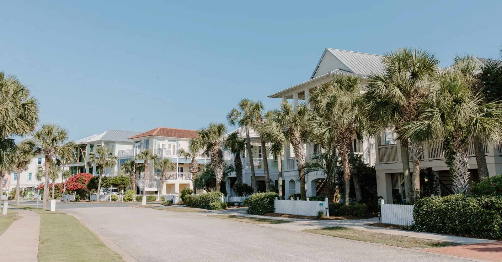 Street of raised Florida homes behind palm trees and white picket fences under a clear sky