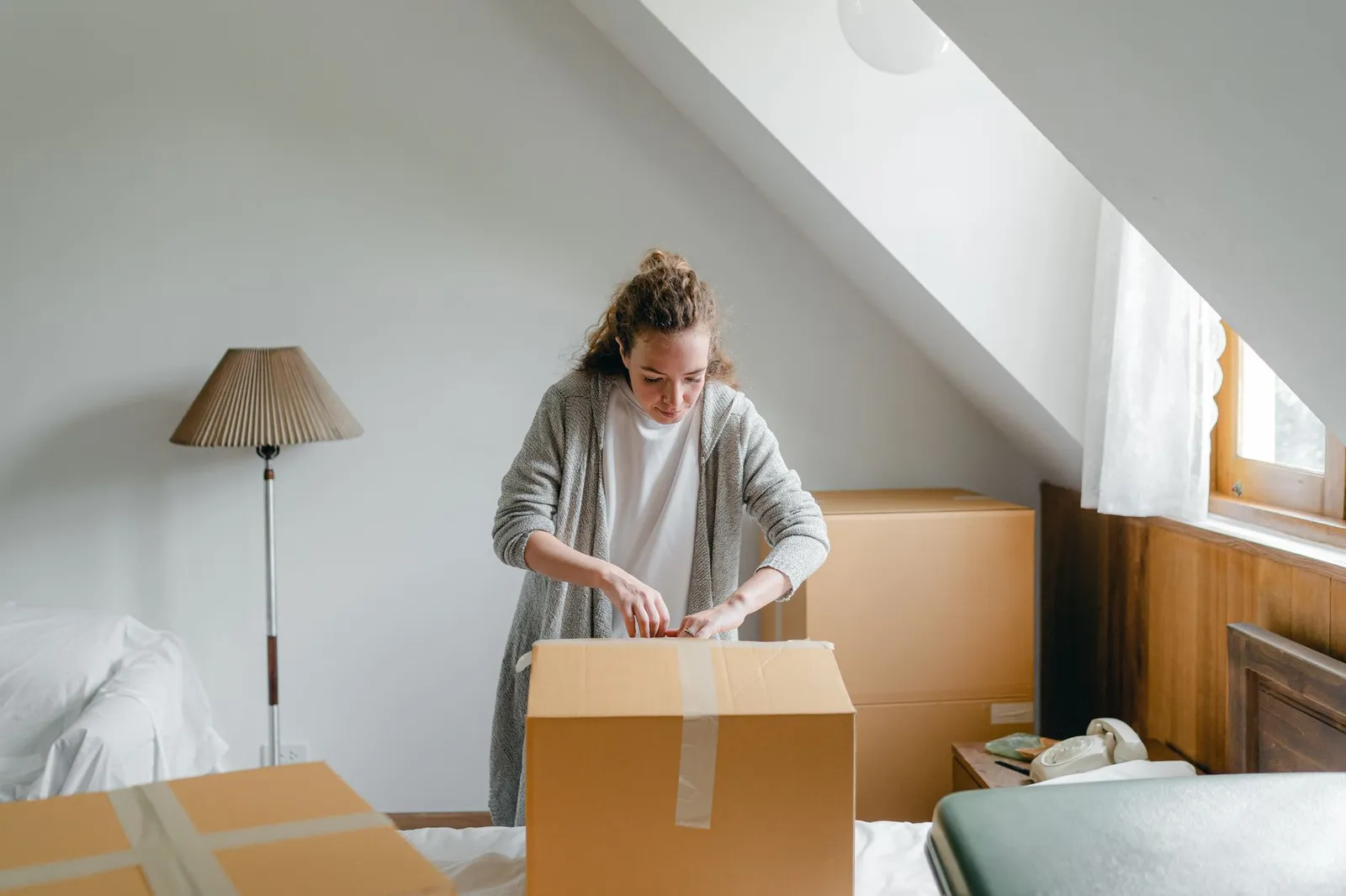 A young woman opening a taped cardboard moving box on a bed in a small attic bedroom