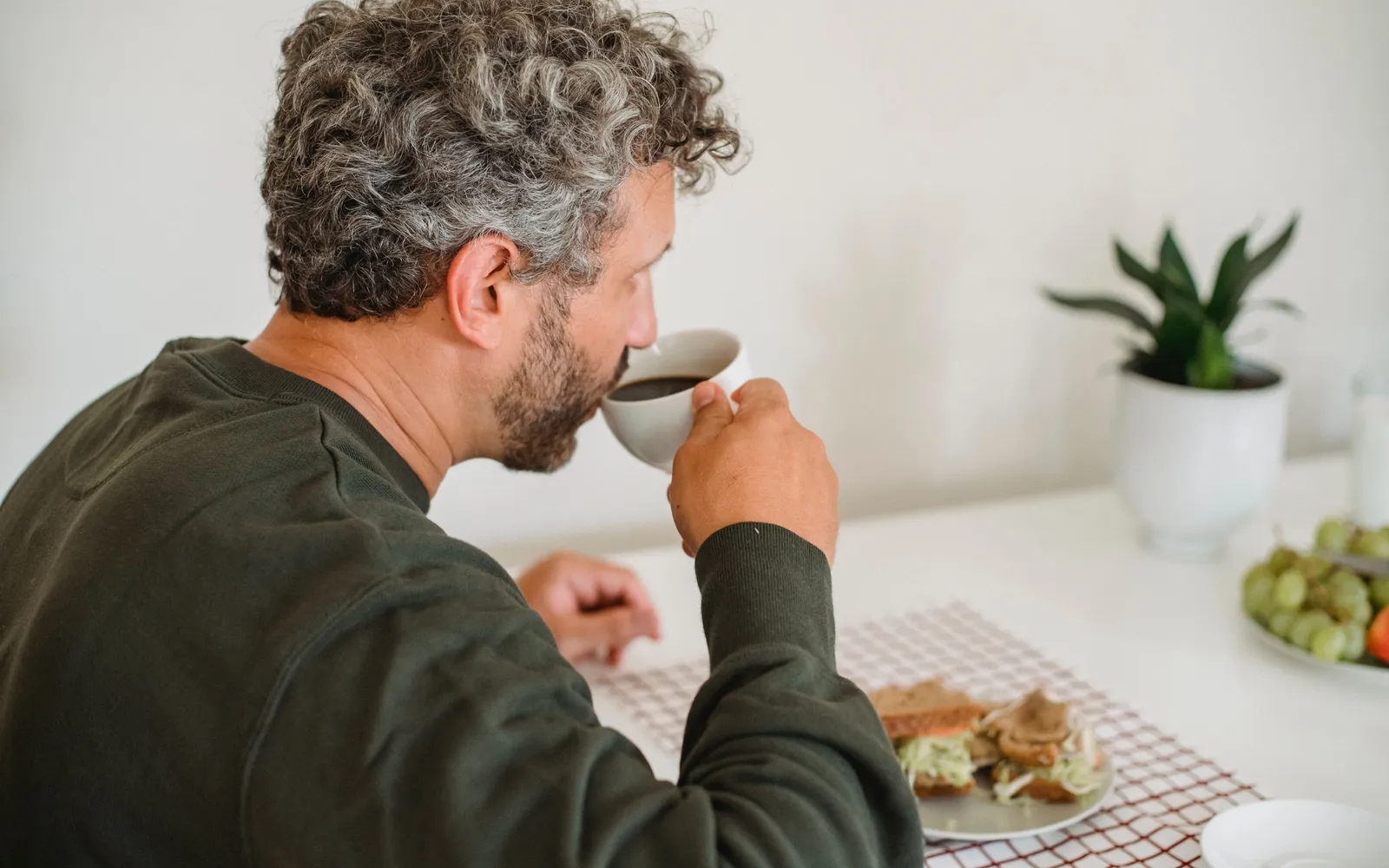 A gray-haired man sips coffee at a white kitchen table beside a sandwich and a potted plant