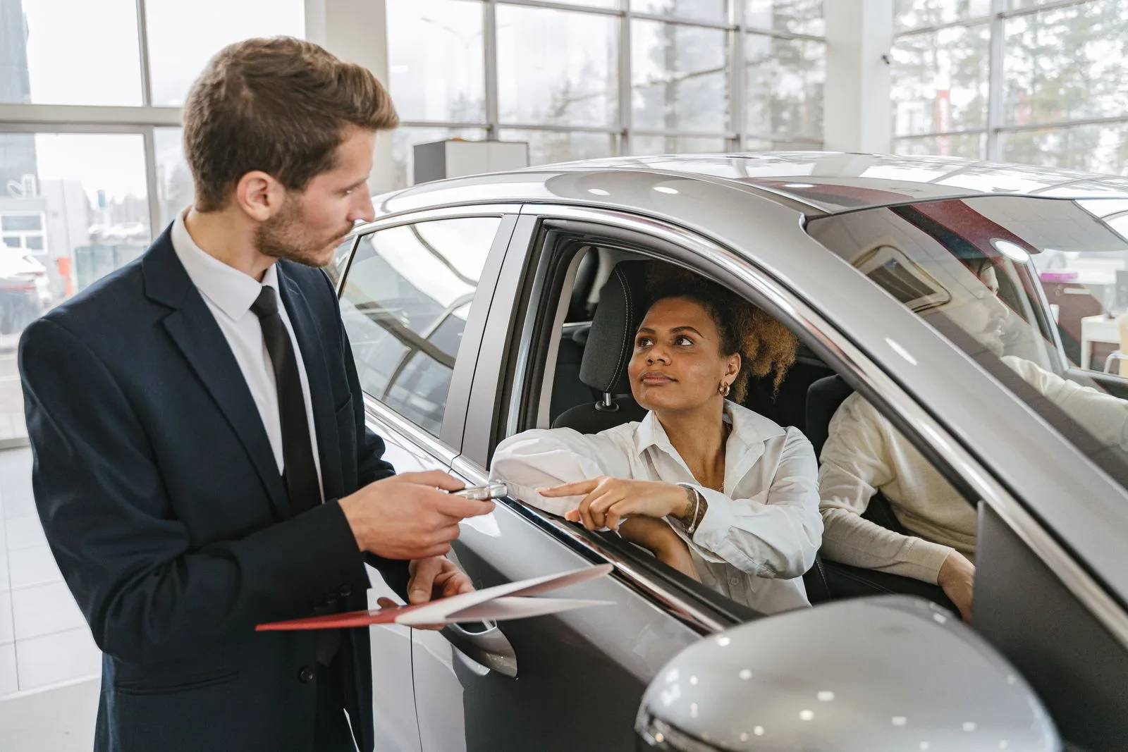 A car dealer hands keys to a woman sitting in a vehicle inside a showroom