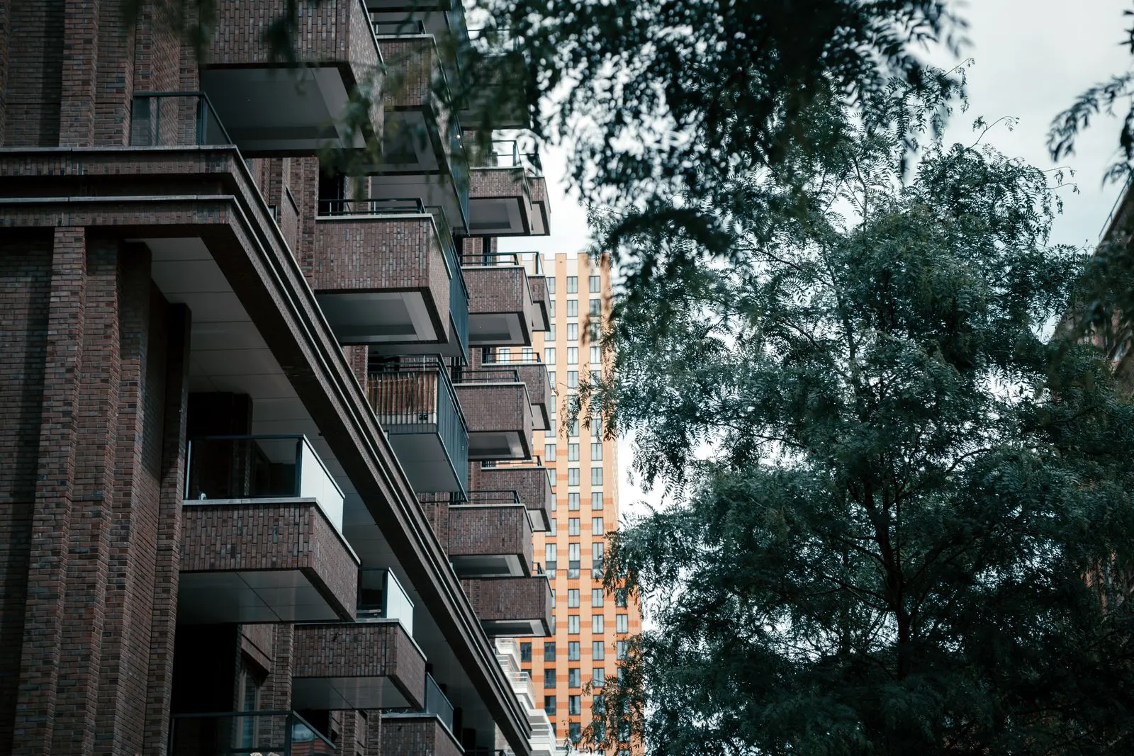 Brick apartment building with stacked balconies behind a leafy tree, a taller residential tower in the background