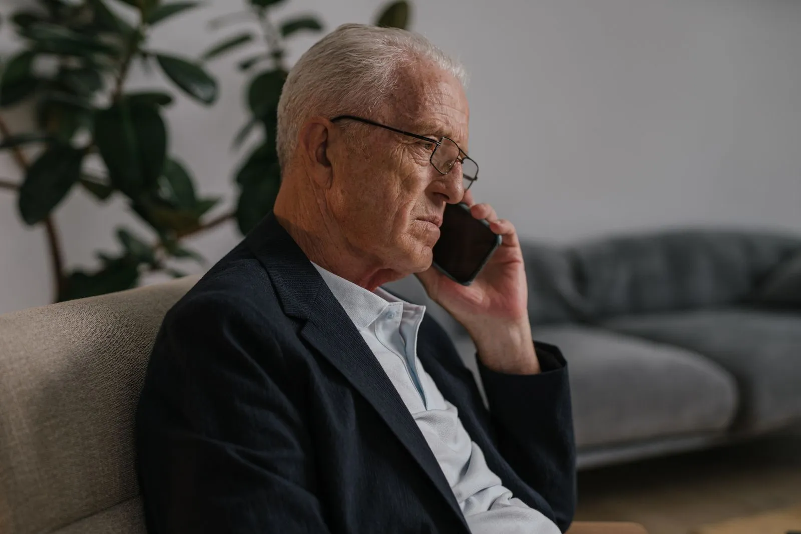Gray-haired man in glasses and a dark blazer on a phone call while seated in a living room