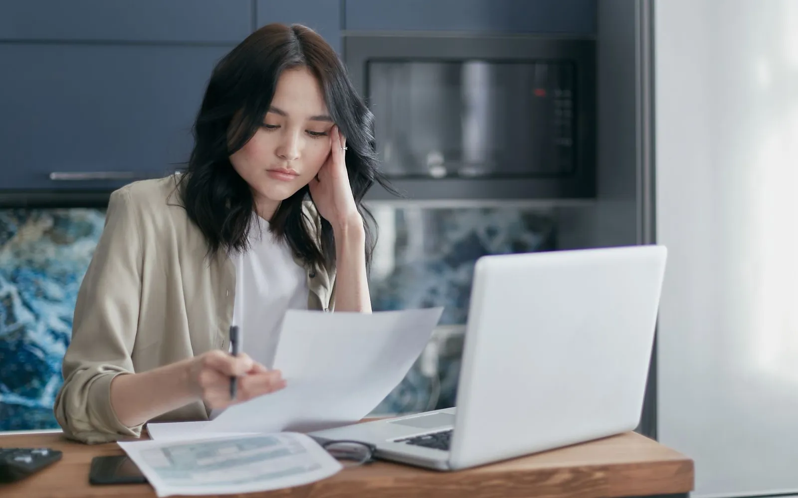 Woman at a kitchen table reading a paper statement next to an open laptop
