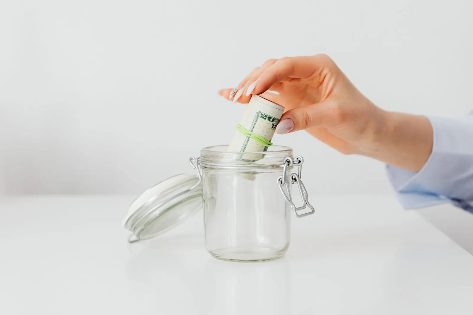 A hand dropping a rolled stack of dollar bills into an open glass jar on a white table