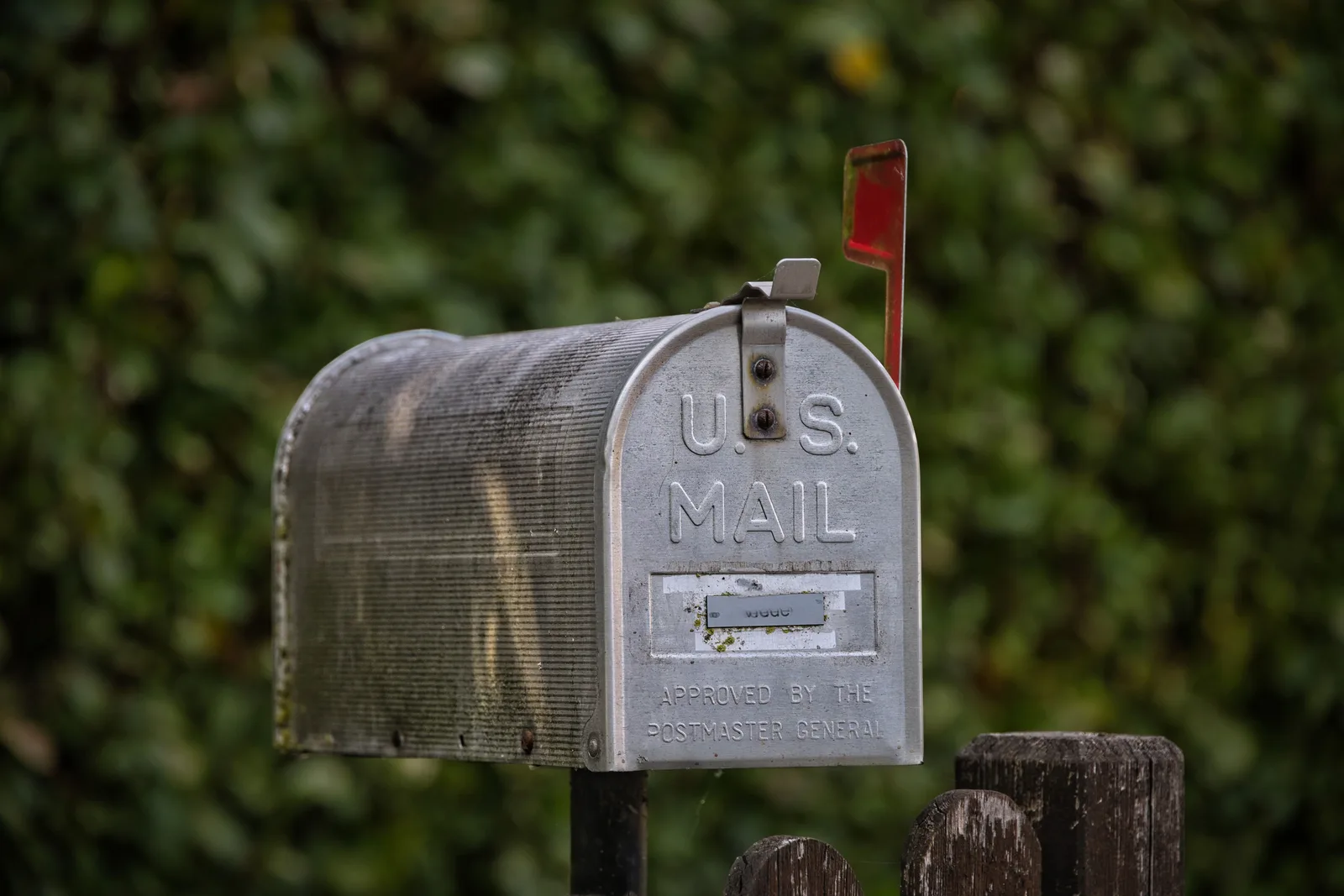 A weathered U.S. Mail curbside mailbox with its red flag raised, against green foliage