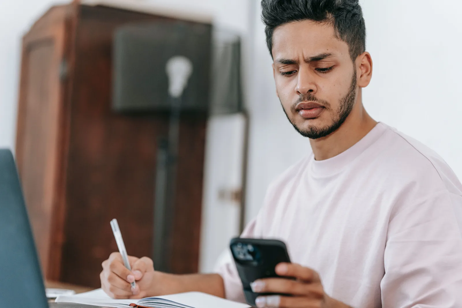 A person at a desk writing in a notebook while holding a mobile phone