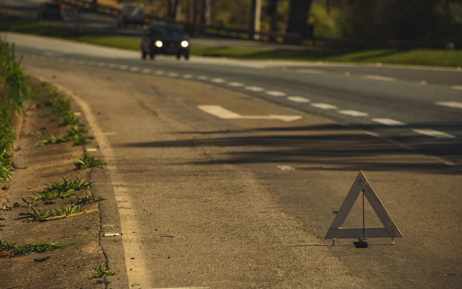 A reflective warning triangle set on the shoulder of a two-lane road as a car approaches with its headlights on