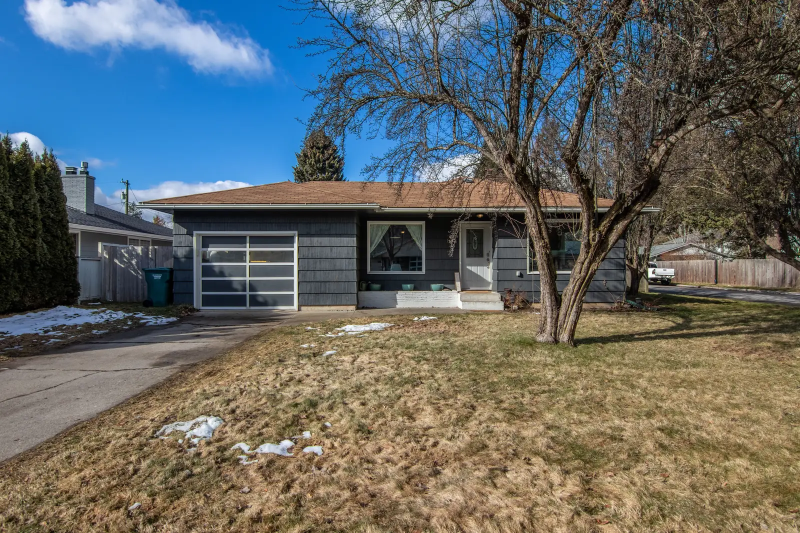 Single-story gray house with a bare tree and a patchy winter lawn under a blue sky