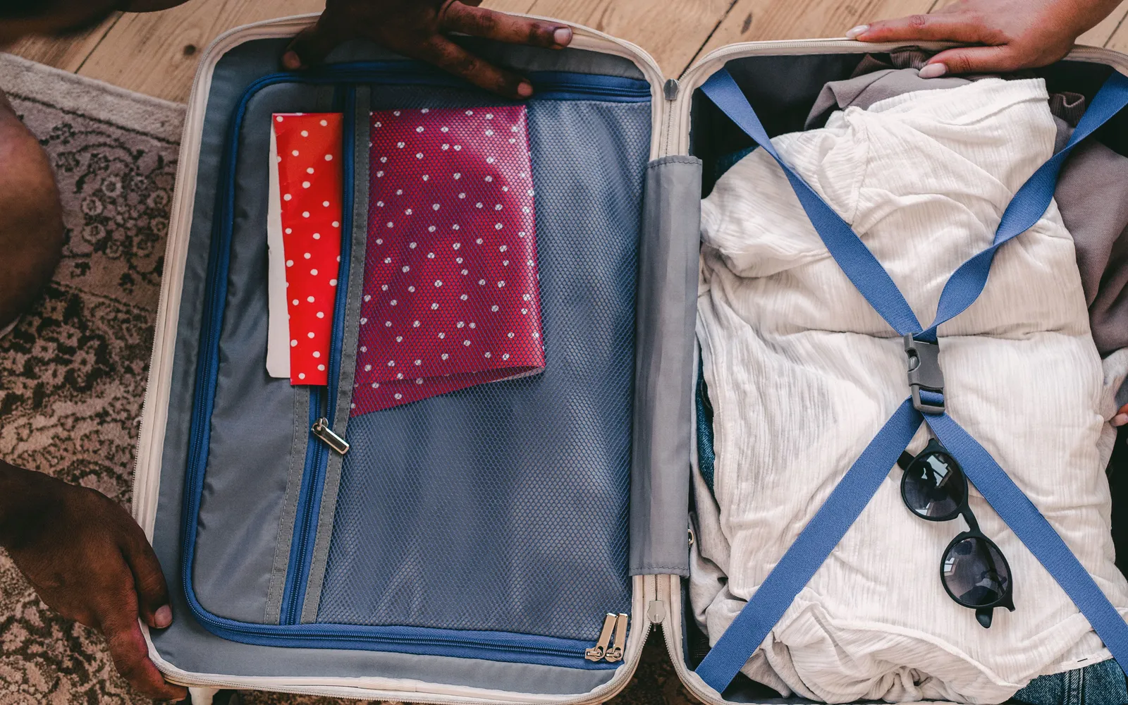 Two people packing an open suitcase on a wooden floor, clothes strapped in on one side and folders in the lid