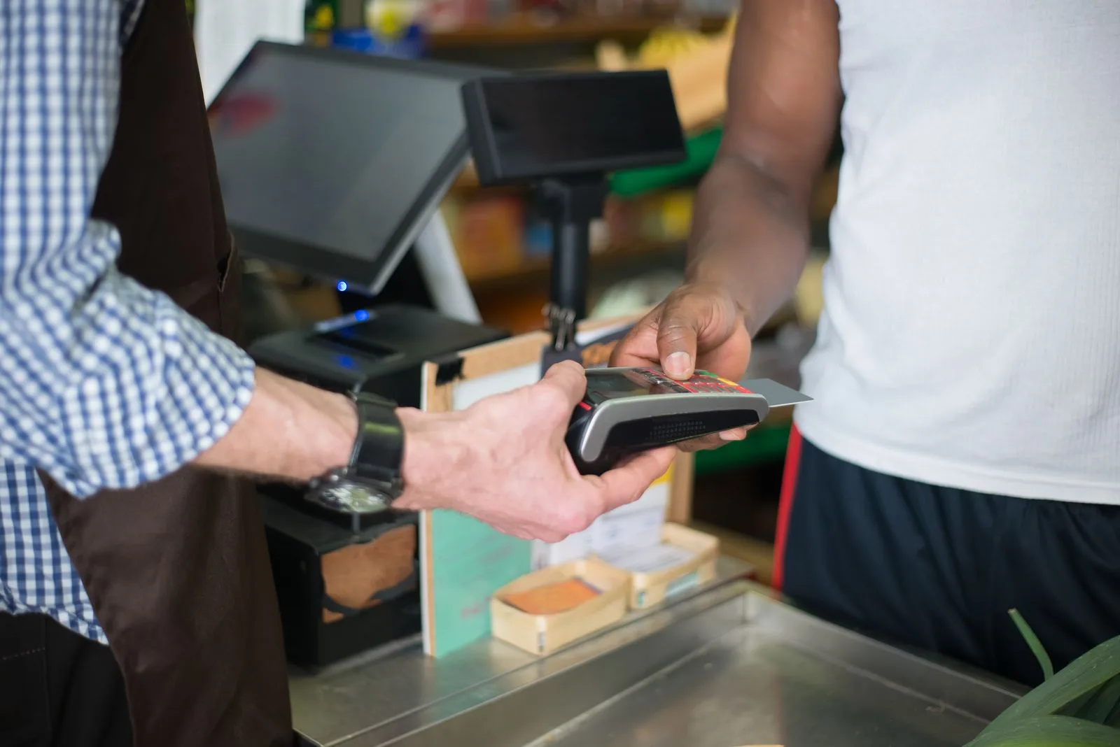 A shopkeeper holds out a handheld card terminal while a customer taps a payment card