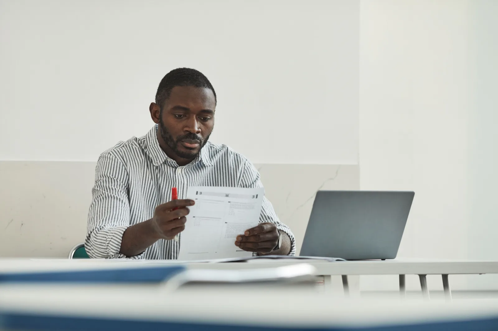 A teacher grading papers at a desk beside a laptop