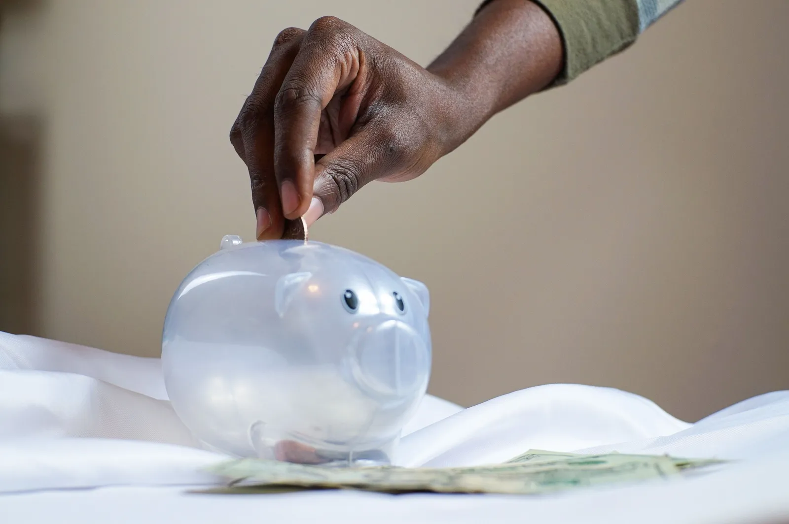 Hand dropping a coin into a clear plastic piggy bank with dollar bills on the table beside it