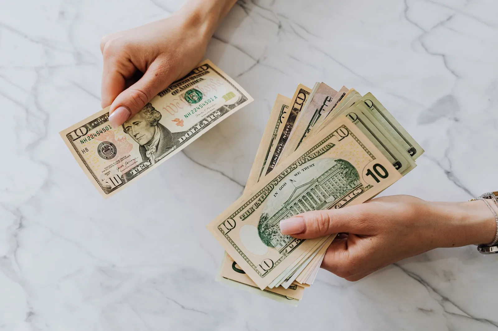 Two hands holding US ten dollar bills over a white marble table