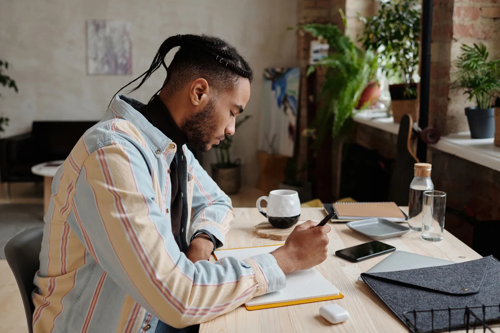 Young man writing figures in a notebook at a wooden desk with a coffee mug and a phone