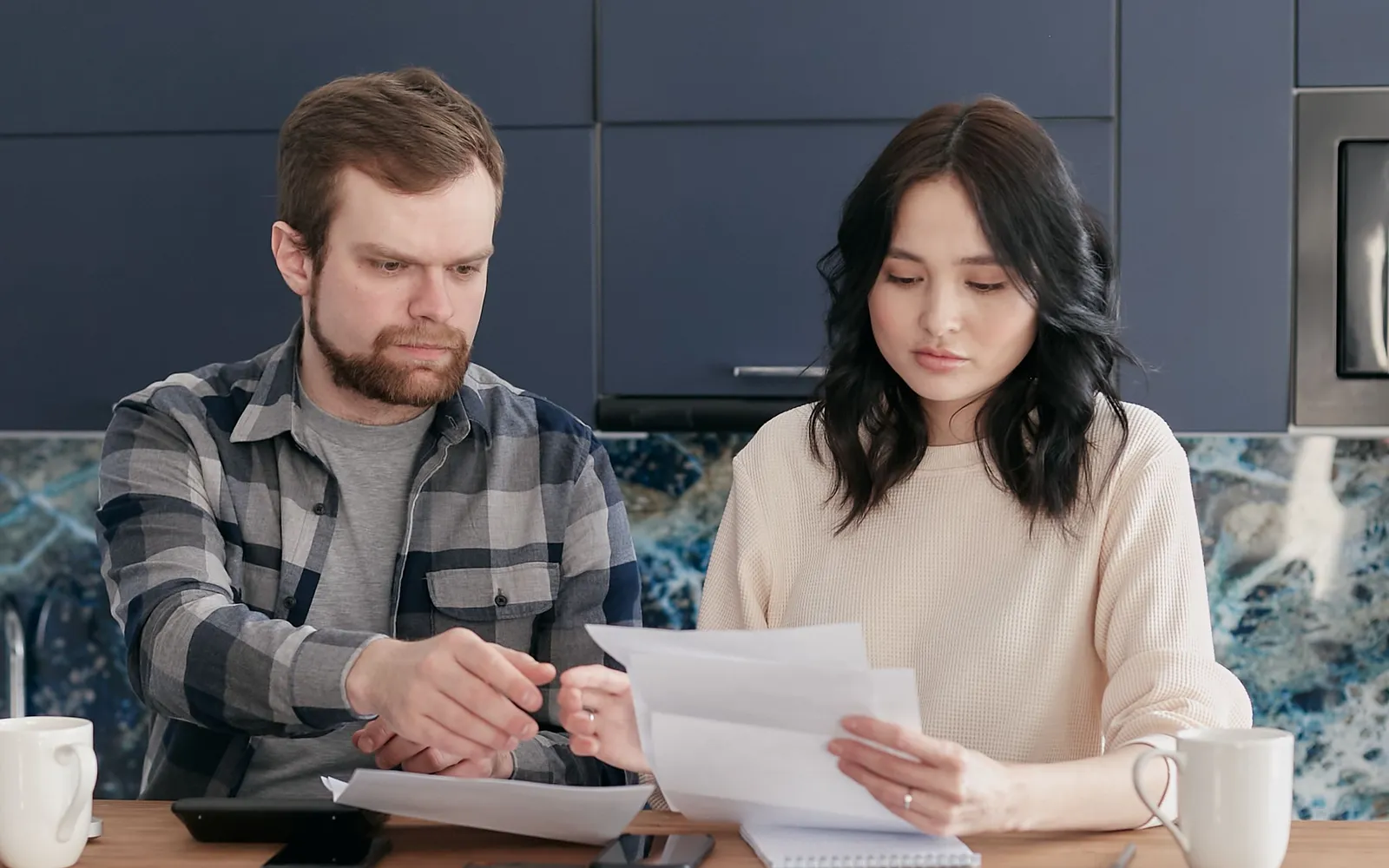 A young couple at a kitchen counter reading through paperwork together, coffee mugs on either side