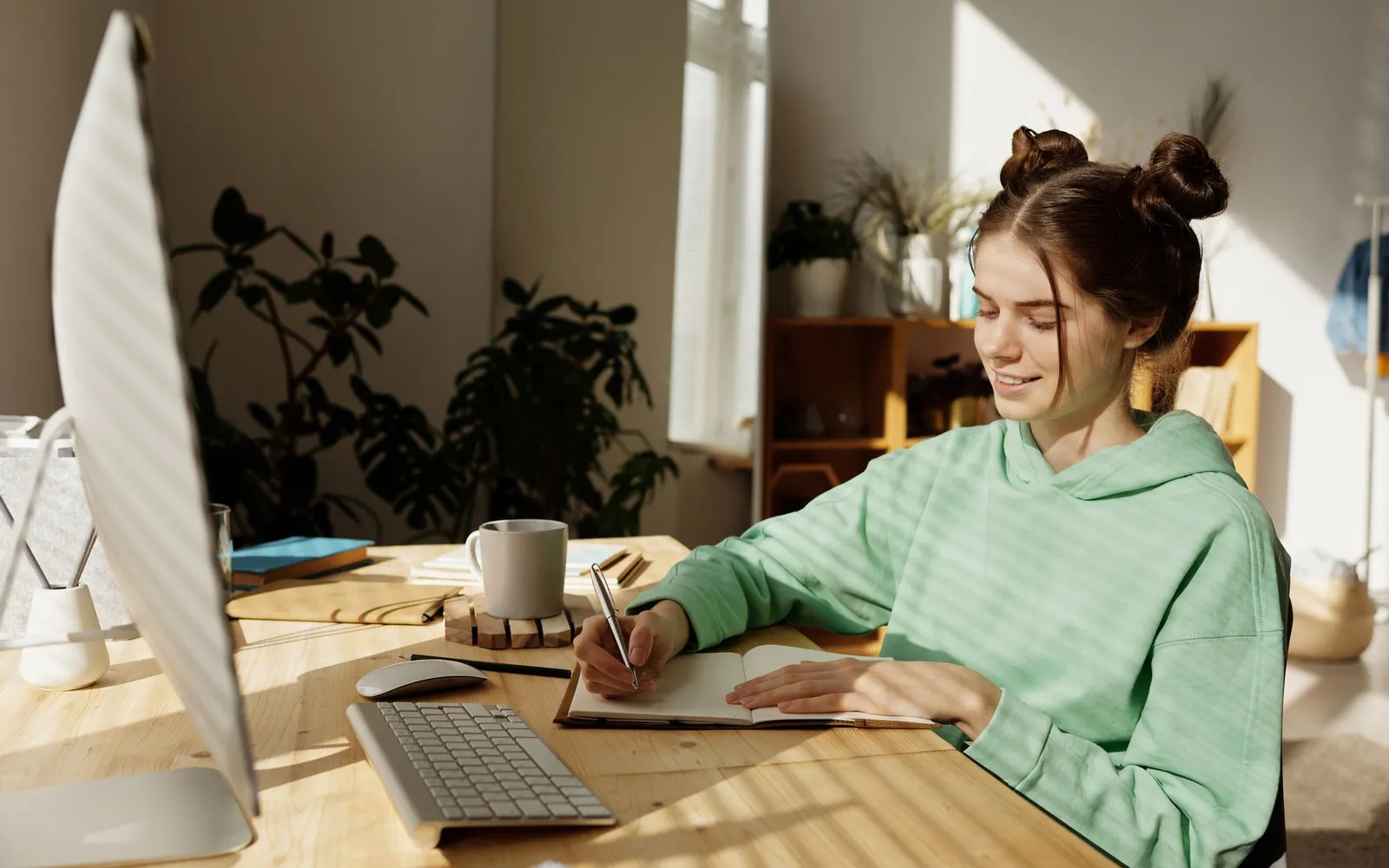 A teenager in a green hoodie writing in a notebook at a sunny desk