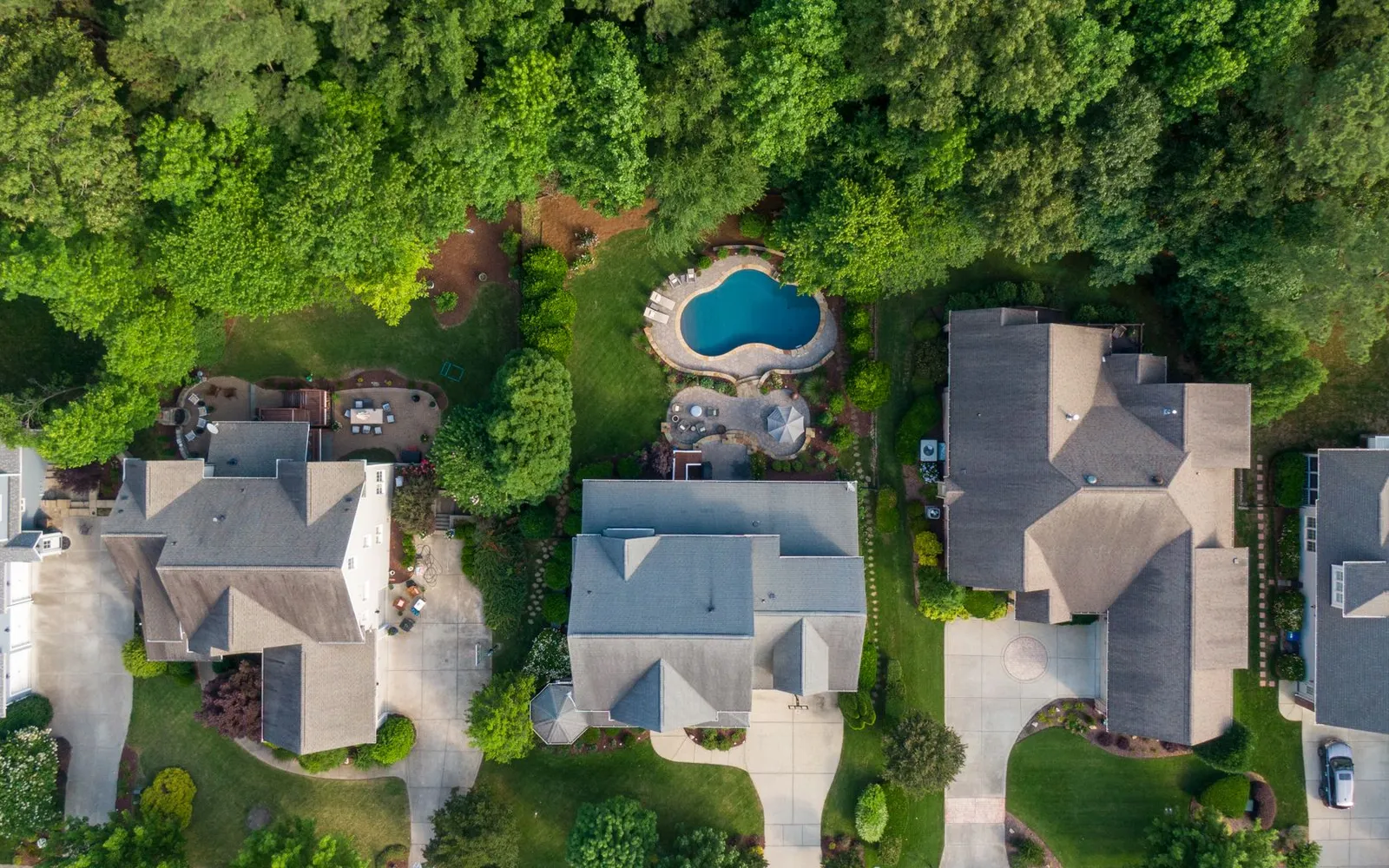 Overhead drone view of suburban rooftops, driveways and a backyard pool surrounded by trees