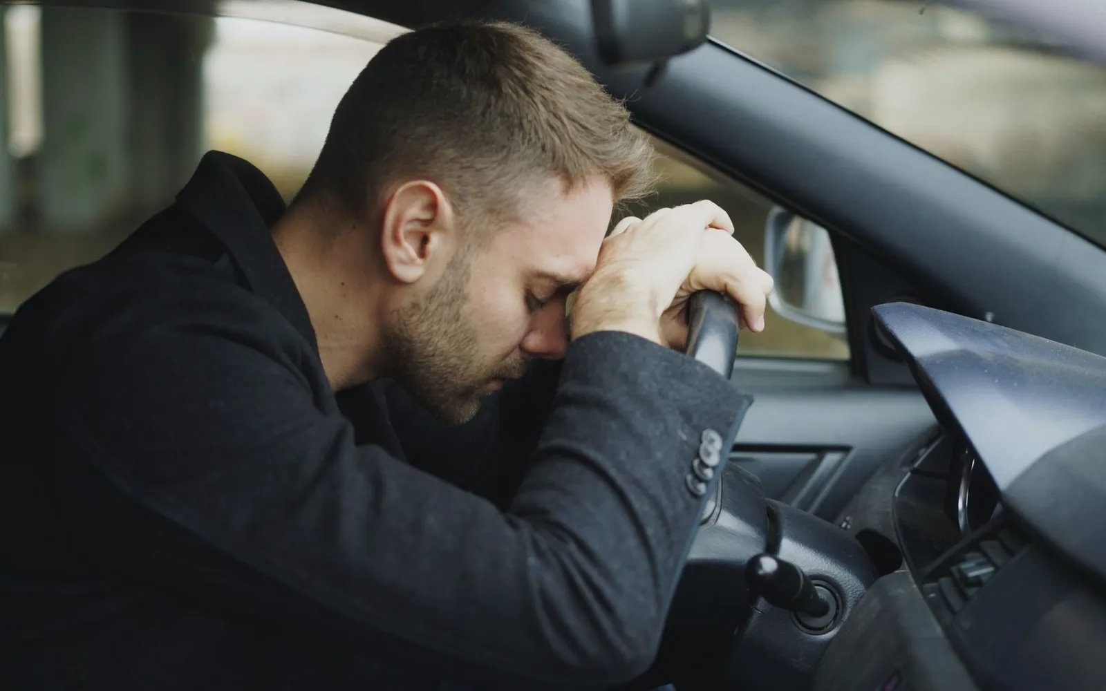 A man in a parked car resting his forehead on his hands on the steering wheel