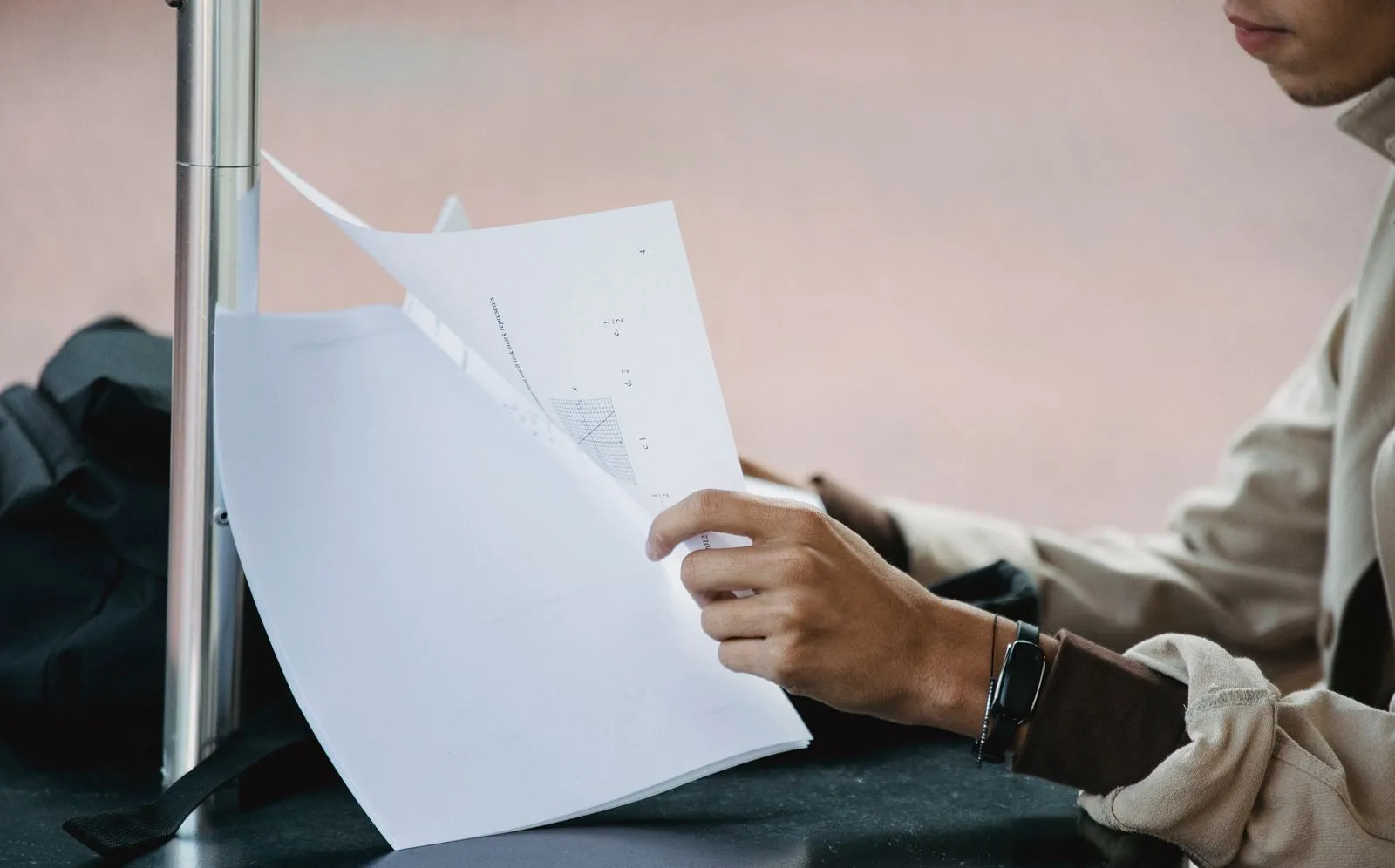A student flipping through pages of coursework at a table with a backpack beside him