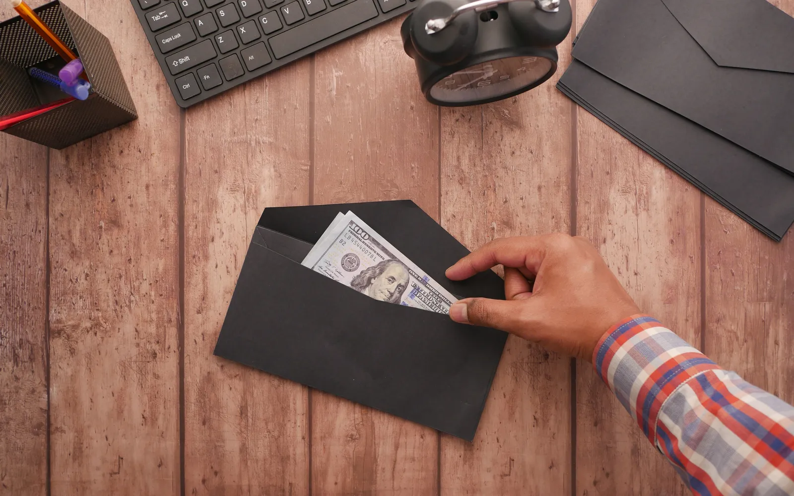 Hand tucking a hundred-dollar bill into a black envelope on a wooden desk beside an alarm clock