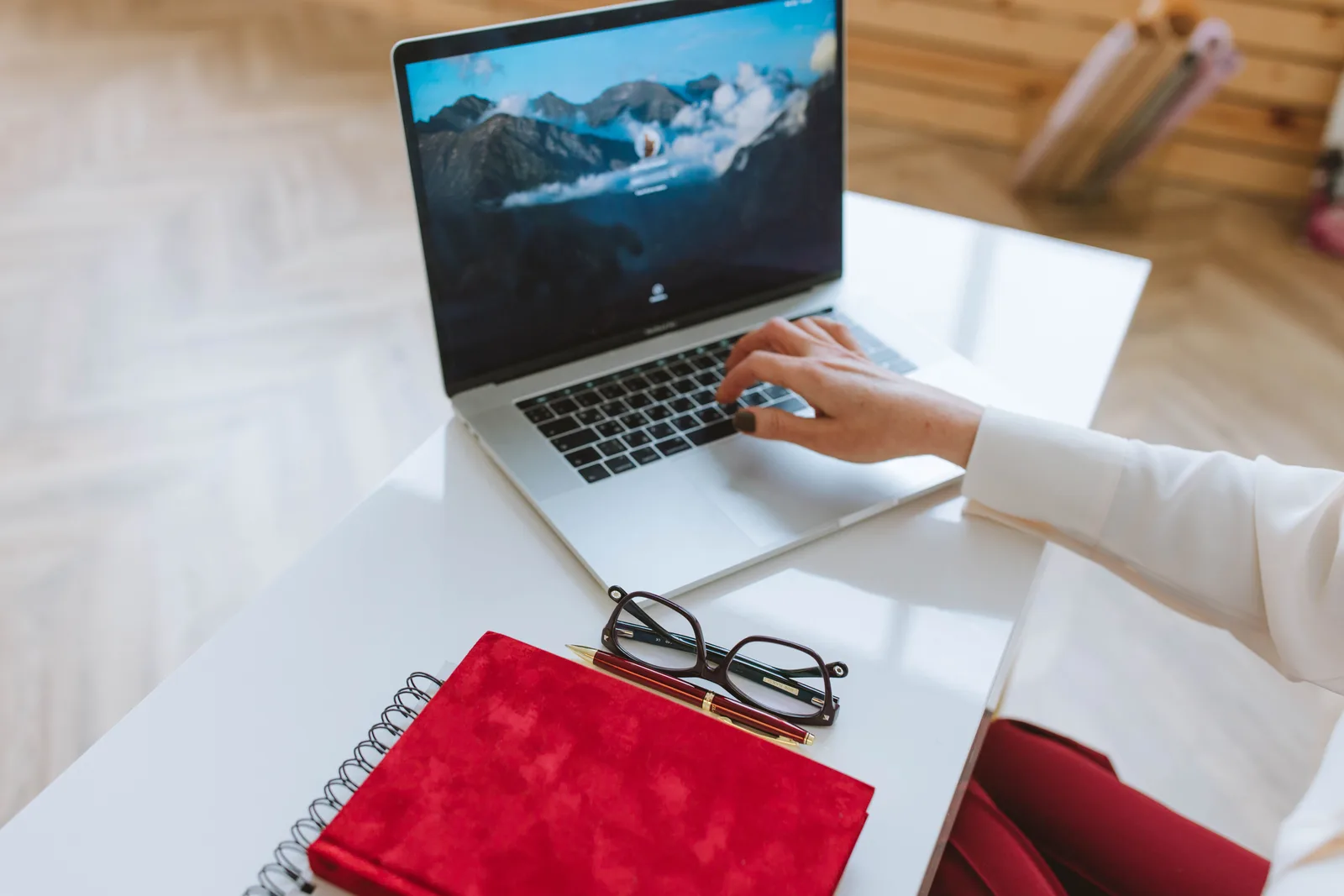 Person working at a laptop with a red notebook and glasses on a white desk