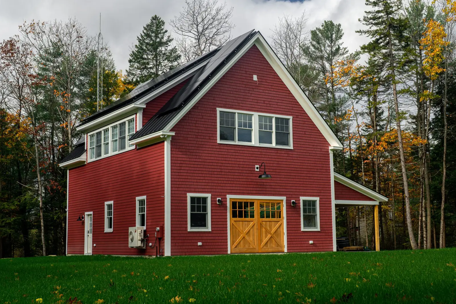 Red clapboard house with black solar panels on its metal roof, surrounded by pines and autumn maples