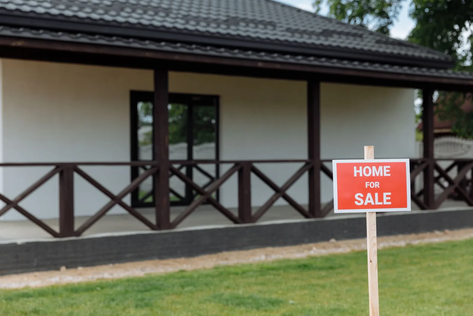 Red Home For Sale sign on a wooden stake in the front lawn of a house with a covered porch