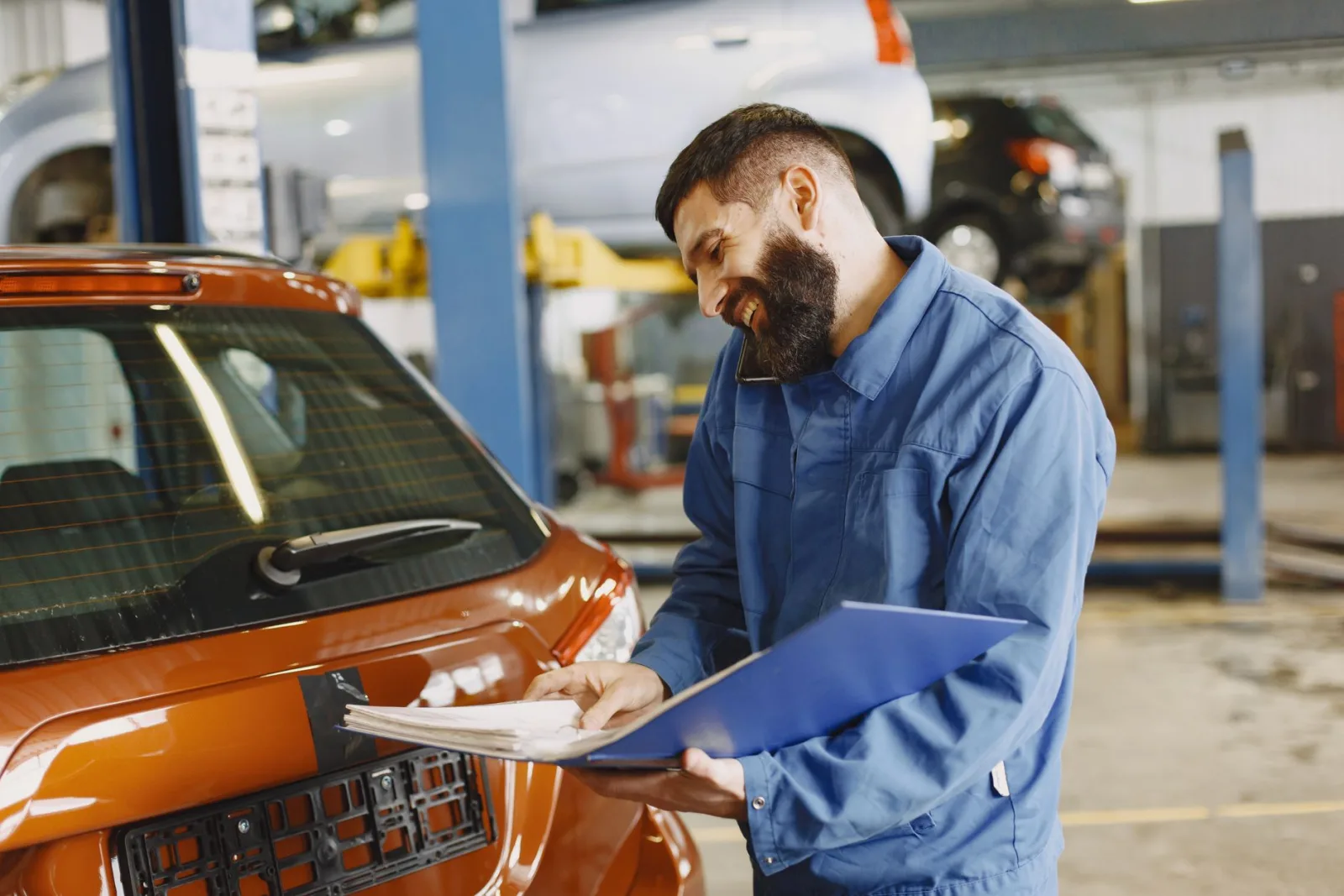 Technician in blue coveralls holding a clipboard of paperwork beside an orange car inside a repair shop