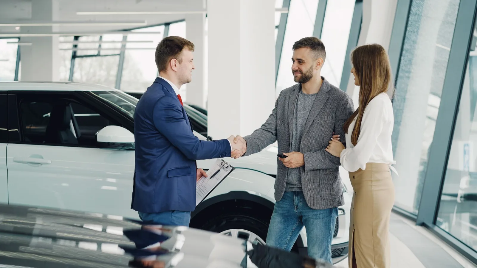 A salesperson shaking hands with a couple beside a white SUV in a car dealership showroom