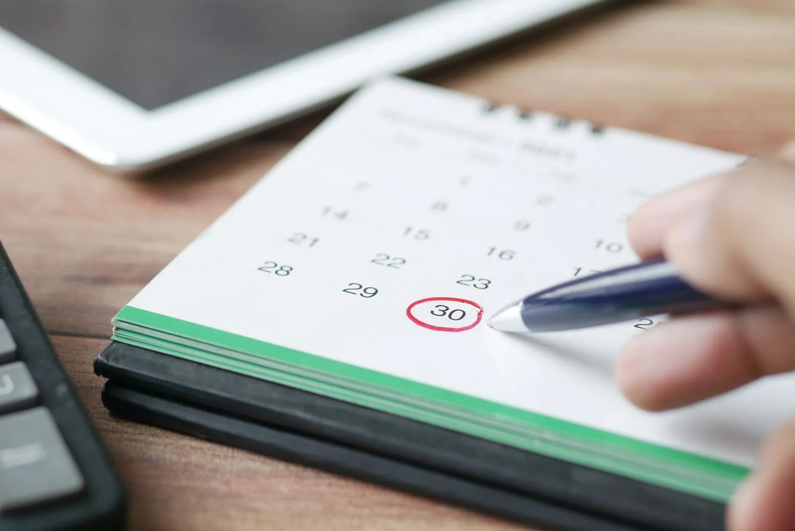 A hand circling a date in red on a desk calendar beside a tablet and a calculator