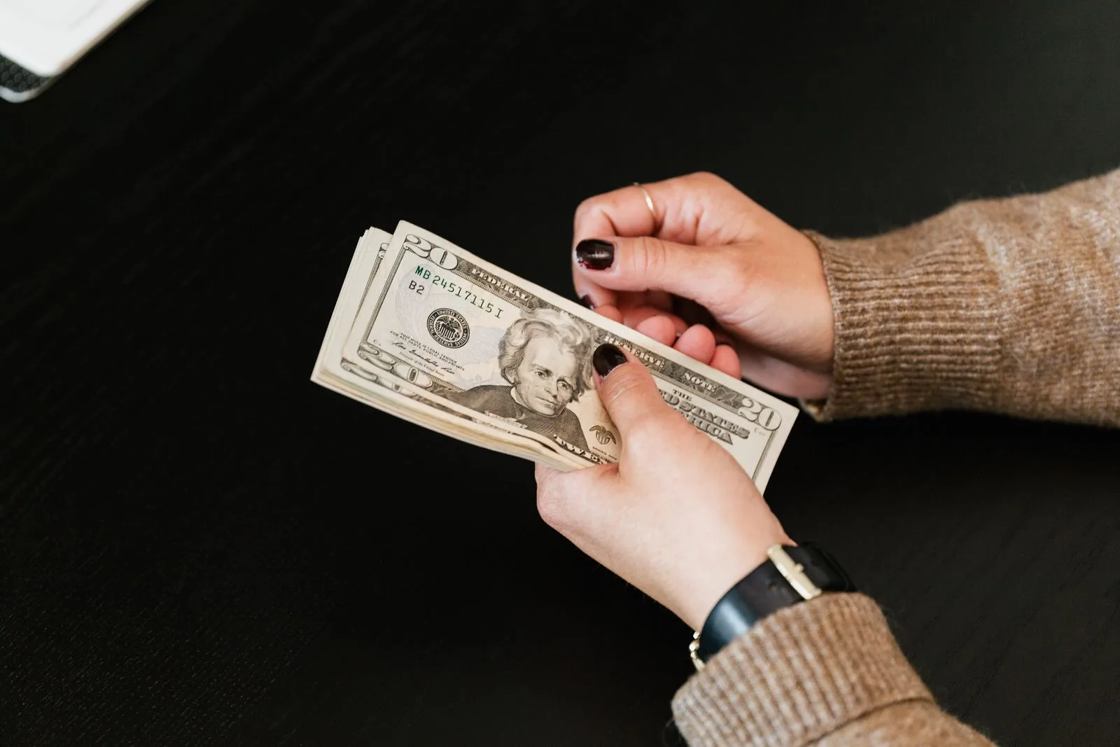 A pair of hands counting a folded stack of twenty dollar bills on a dark table